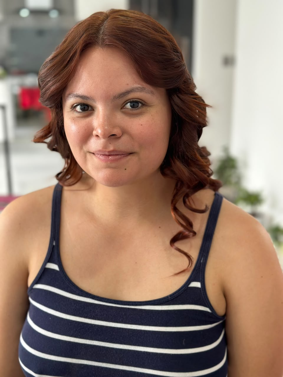 Antes de maquillaje social: rostro sin acabado de evento, cabello rojizo con ondas suaves y top a rayas.