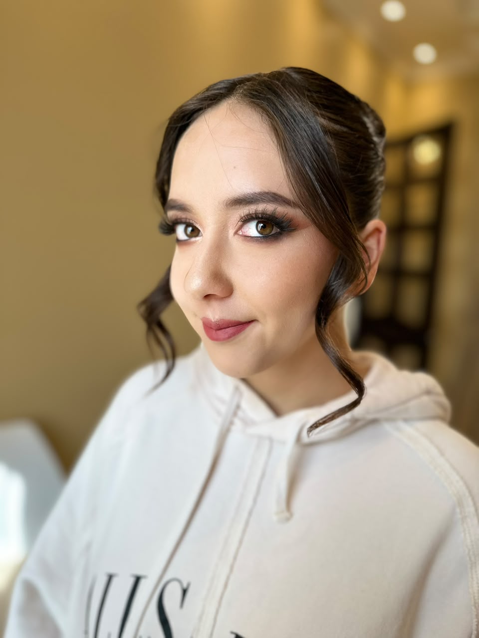 Maquillaje glam con piel luminosa, mirada intensa y peinado recogido con mechones frontales.